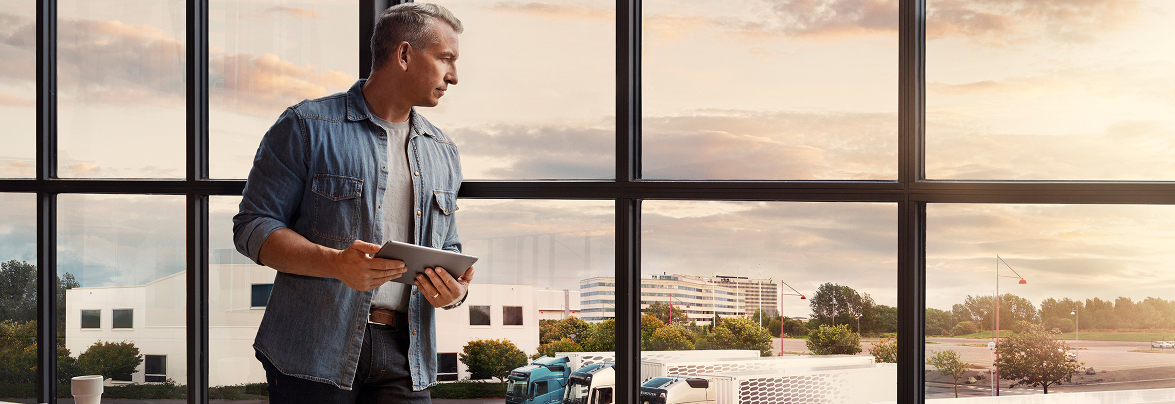 Um homem segurando um tablet fica perto de uma janela e olha para sua frota de caminhões